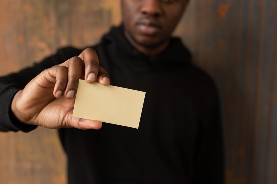 Unrecognizable African American male in casual clothes demonstrating yellow mock up business card in hand while standing near wooden wall